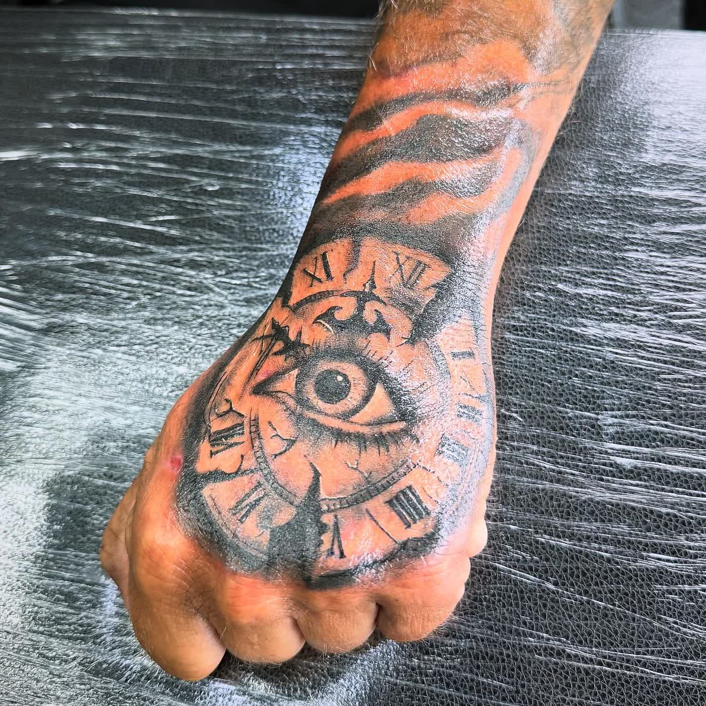 A clock and eye tattooed in black and grey across the back of a hand