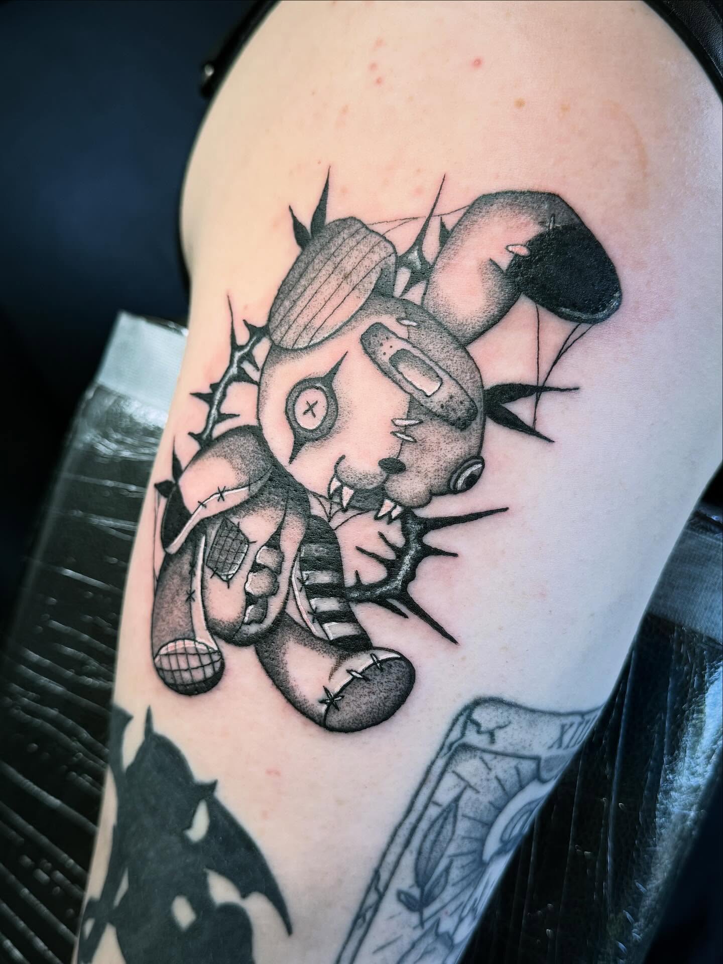 A stitched and patched teddy bear tattooed in fine black and grey shading